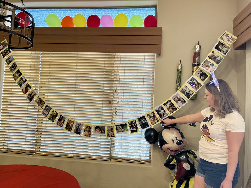 Mom hangs a photo banner showing memories from her daughter’s first three years at a Mickey Mouse birthday party