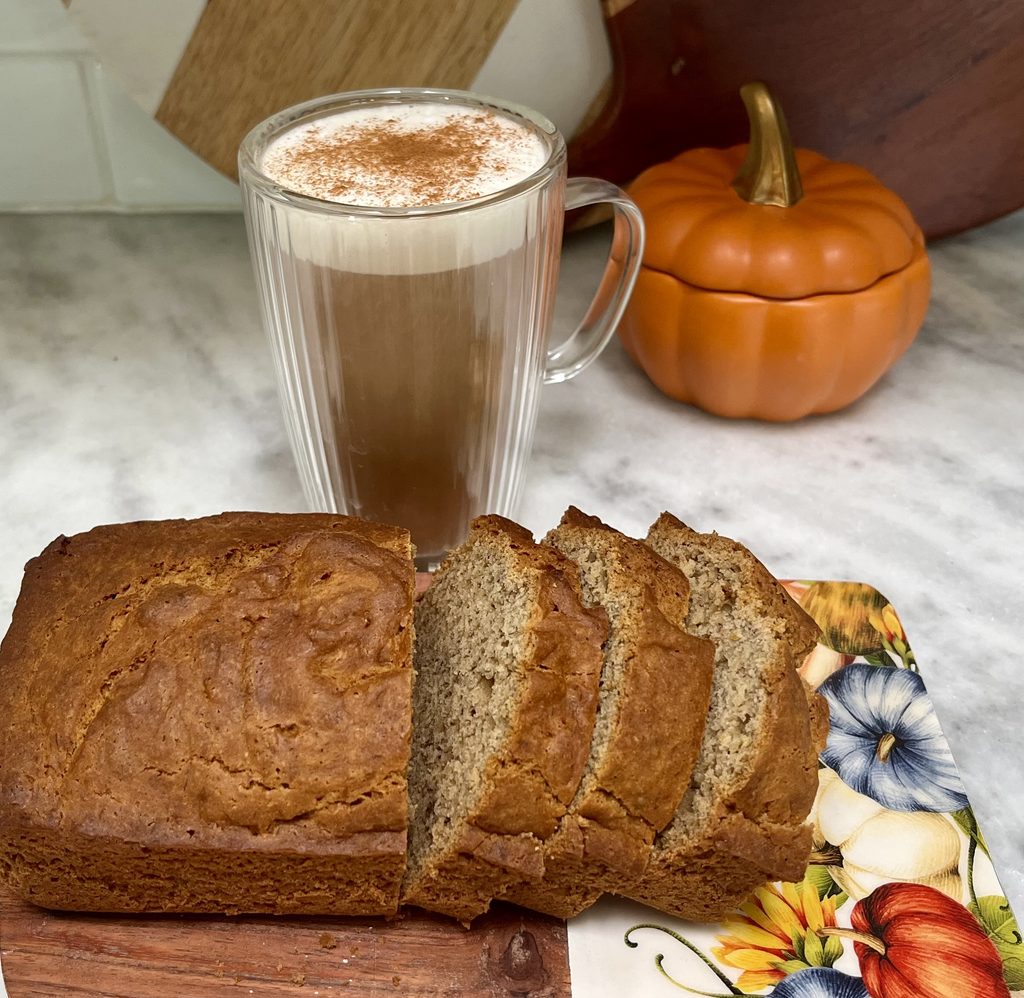 Sliced homemade gluten-free banana bread beside a cinnamon-topped hot drink and a small pumpkin decoration.
